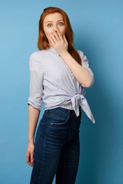 Beline bağlanmış çizgili tişörtlü kızıl saçlı kız mavi arka planda duruyor ve gözlerini kocaman açıyor. Ağzını eliyle kapatıyor.