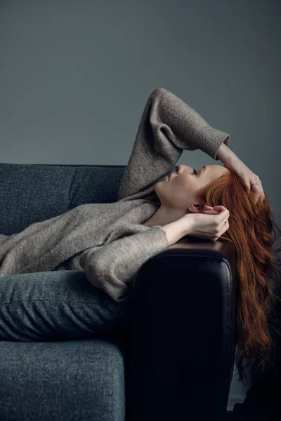 Uma menina ruiva em um casaco bege jaz no sofá, inclinando a cabeça para trás no braço, passando a mão pelo cabelo . — Fotografia de Stock