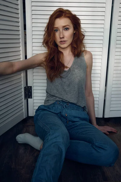 Menina de cabelos vermelhos em uma camiseta e calças senta-se no chão contra uma tela branca e olha para a câmera — Fotografia de Stock