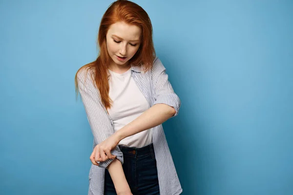 Chica pelirroja con una camisa a rayas se levanta sobre un fondo azul y se sube la manga, mirando sus manos . — Foto de Stock