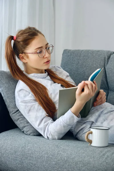 Röd tjej i pyjamas och runda glasögon ligger på soffan och läser en bok. Stockbild