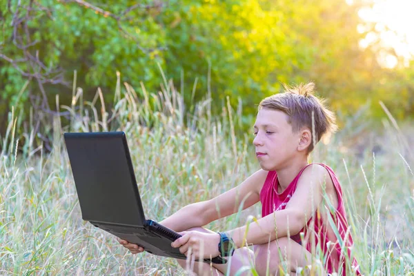 A boy with a laptop in a clearing in the forest is engaged in work and play outdoors in nature