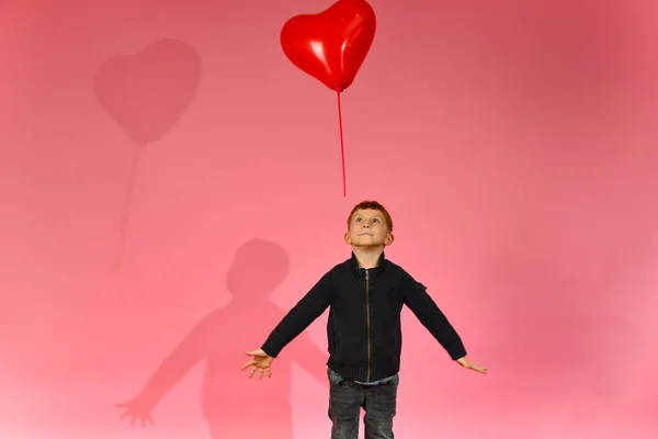 一个男孩在工作室的粉红色背景上投掷并捕捉到一个心形气球 — 图库照片