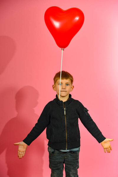 A boy throws and catches a heart-shaped balloon on a pink background in the studio.