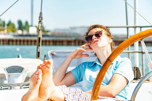 A girl in glasses sits near the wheel next to on the deck of a yacht looking around