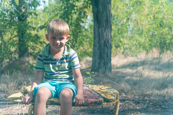 男孩坐在一张旧板凳上 在一个废弃的公园里 带着悲伤的表情环顾四周 — 图库照片