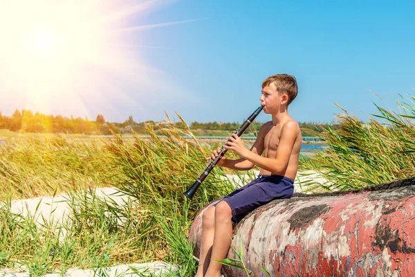一个男孩坐在海边的一艘旧木船上 坐在一条黑色的单簧管上 望着旁边 从侧面看风景 — 图库照片
