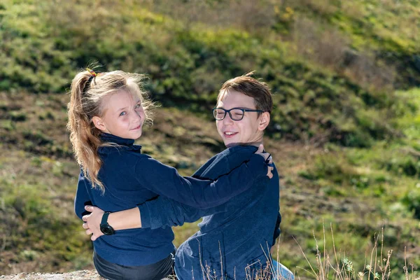 A boy with a girl are sitting on a cliff, two friends, a brother and sister are sitting in an embrace and look into the camera through the back
