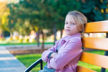 Bir kız pembe bir ceket ön ve parkta sarı bir bankta oturmuş kameraya bakarak onun kollarında katlanmış