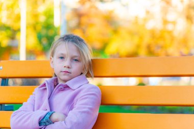 Bir kız pembe bir ceket ön ve parkta sarı bir bankta oturmuş kameraya bakarak onun kollarında katlanmış