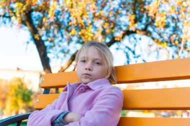Bir kız pembe bir ceket ön ve parkta sarı bir bankta oturmuş kameraya bakarak onun kollarında katlanmış