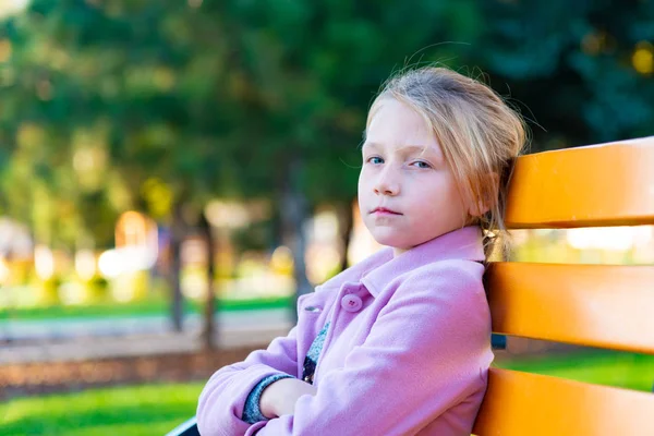 Bir kız pembe bir ceket ön ve parkta sarı bir bankta oturmuş kameraya bakarak onun kollarında katlanmış