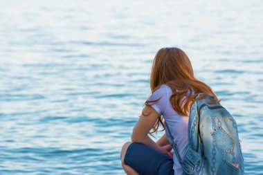 Bir kız Kızıl saçlı ve bir sırt çantası bir taş deniz tarafından ele geçirilmesi ve mesafe içine bakan.