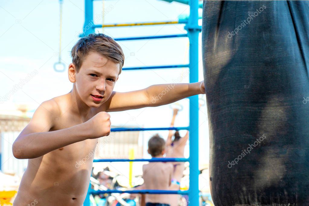 A boy with a furious and angry face waving his hand and punching a pear, looking into the camera. Is engaged in boxing on the beach, on the beach