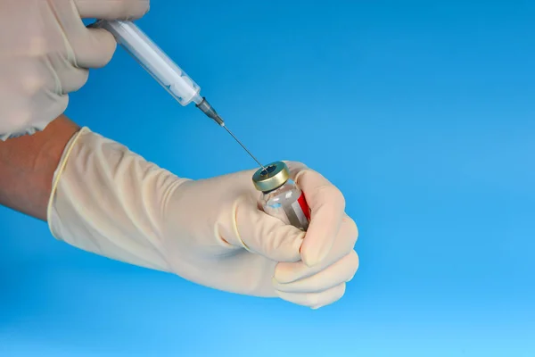 En lege i medisinske hansker tar medisin fra et hetteglass på blå bakgrunn inn i en sprøyte – stockfoto