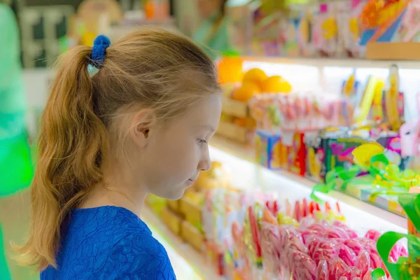 Uma menina fica na janela da loja e olha para doces e doces . — Fotografia de Stock