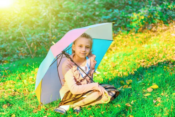 Девушка сидит на зеленой траве с цветным зонтиком, закрываясь от солнечных лучей и летней жары . — стоковое фото