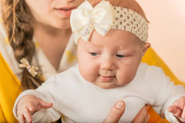 Ein kleines neugeborenes Mädchen mit einer Schleife auf dem Kopf sitzt neben ihrer Mutter auf dem Arm. — Stockfoto