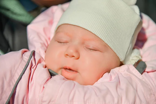 A newborn baby in a pink jacket and white hat is sleeping on the street in a stroller