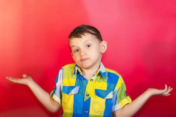Un niño con una camisa de color se encoge de hombros y extiende los brazos hacia un lado, sobre un fondo rojo — Foto de Stock