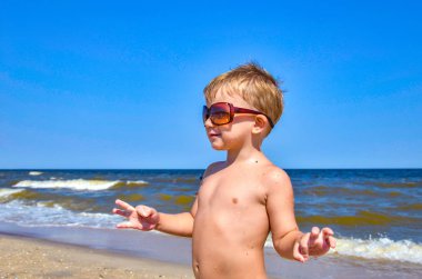 Gözlüklü bir çocuk deniz kenarında sahilde duruyor.