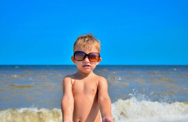 Gözlüklü bir çocuk deniz kenarında sahilde duruyor.