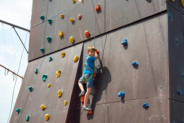 Chłopiec wspina się na szczyt sztucznej ściany w skrajnym parku, trzymając na liny bezpieczeństwa. — Zdjęcie stockowe