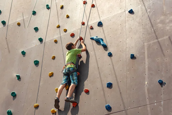 Chłopiec wspina się na szczyt sztucznej ściany w skrajnym parku, trzymając na liny bezpieczeństwa. — Zdjęcie stockowe