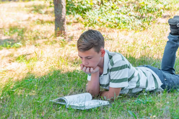 El niño se encuentra en el parque en la hierba y está estudiando en la naturaleza, la educación y la ciencia, el libro está acostado en la hierba . — Foto de Stock