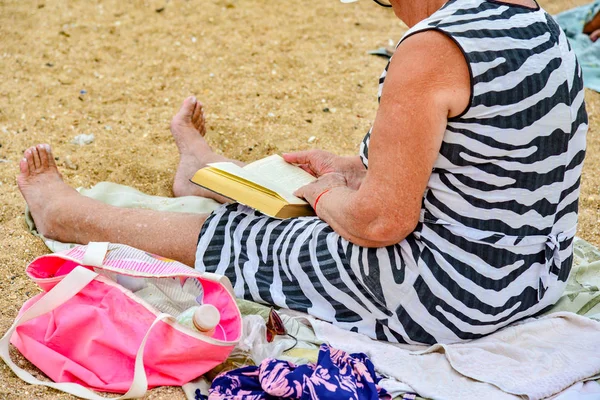 La abuela lee un libro en la playa sentado en la arena junto al mar . — Foto de Stock