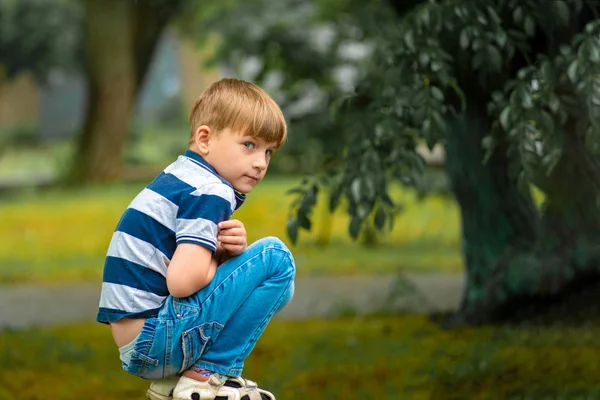 Um menino de jeans azul agacha-se no parque olhando em volta . — Fotografia de Stock