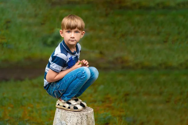 Um menino de jeans azul agacha-se no parque olhando em volta . — Fotografia de Stock