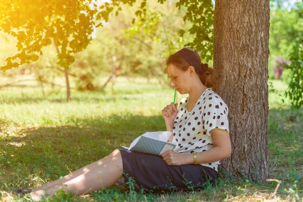 Egy lány egy notebook alatt egy fa, elgondolkodva tartja a ceruzát az ajkak közelében. — Stock Fotó