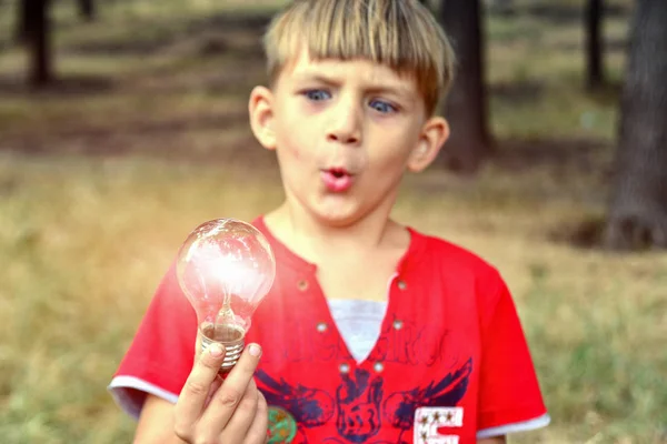 O menino segura uma lâmpada incandescente, que queima em sua mão e olha para ela com surpresa e admiração . — Fotografia de Stock