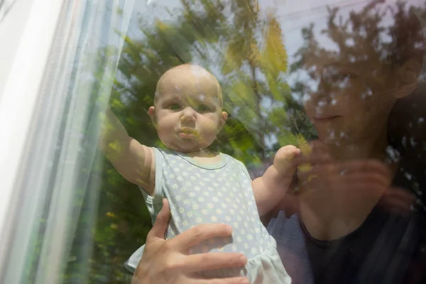 Çocuk pencereden dışarı bakar, küçük kız pencereden dışarı bakar, ve annesi onu tutar. — Stok fotoğraf