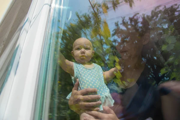 A criança olha pela janela, a menina olha pela janela, e sua mãe a segura . — Fotografia de Stock