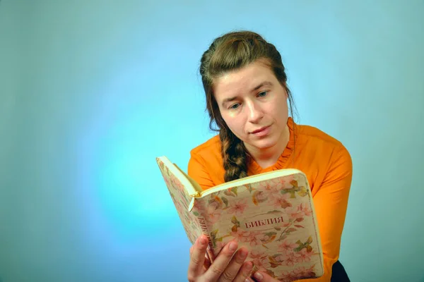 Une fille lit la Bible assise sur une chaise sur un fond bleu. L'inscription en russe - "Bible" sur le livre . — Photo