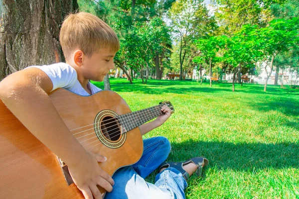 Un niño con una guitarra se sienta debajo de un árbol, foto de gran angular . —  Fotos de Stock