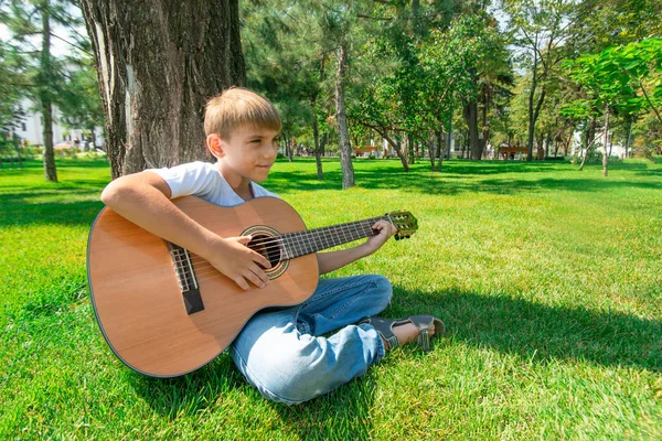 En pojke med en gitarr sitter under ett träd, vidvinkel foto. — Stockfoto
