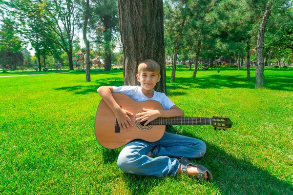 En pojke med en gitarr sitter under ett träd, sjunger sånger och njuter av naturen. — Stockfoto