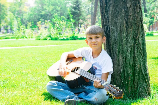 En pojke med en gitarr sitter under ett träd, sjunger sånger och njuter av naturen. — Stockfoto