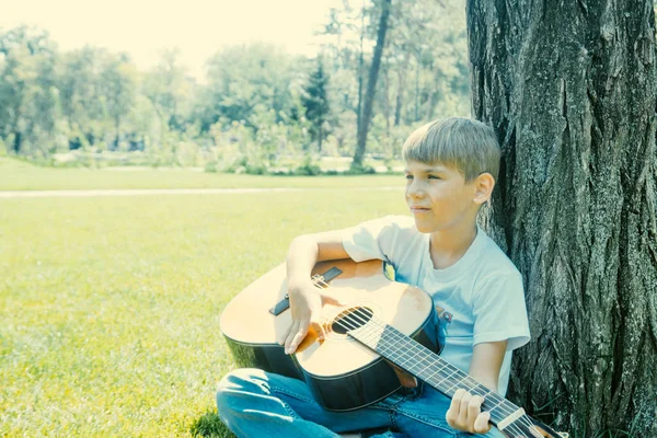 Un chico con una guitarra se sienta bajo un árbol, canta canciones y disfruta de la naturaleza . —  Fotos de Stock