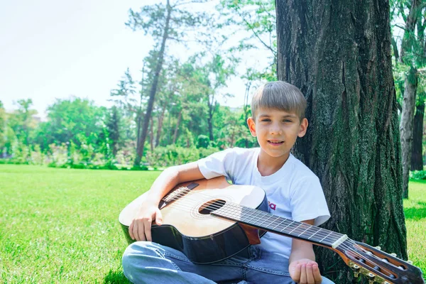 En pojke med en gitarr sitter under ett träd, sjunger sånger och njuter av naturen. — Stockfoto