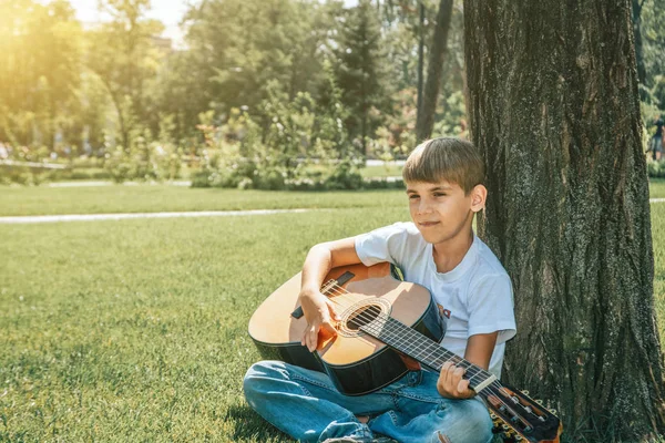 Un chico con una guitarra se sienta bajo un árbol, canta canciones y disfruta de la naturaleza . —  Fotos de Stock