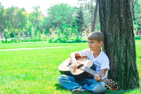 Een jongen met een gitaar zit onder een boom, zingt liederen en geniet van de natuur. — Stockfoto