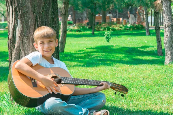 En pojke med en gitarr sitter under ett träd, sjunger sånger och njuter av naturen. — Stockfoto