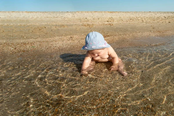 Klein meisje in een hoed zit aan de kust in het water op het strand. — Stockfoto