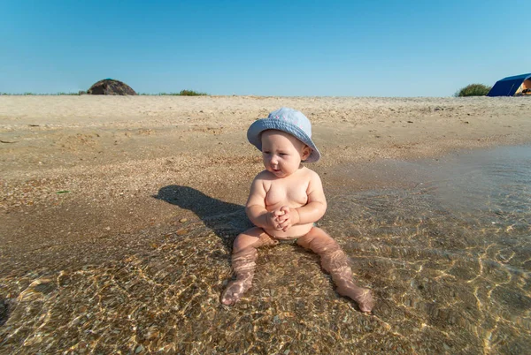 Mała dziewczynka w kapeluszu siedzi na brzegu morza w wodzie na plaży. — Zdjęcie stockowe