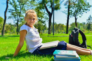 Bir kız ders kitaplarında ödevleri öğretir ve parkta yeşil çim oturur.