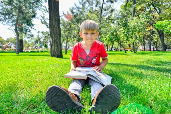 Kırmızı elbiseli bir çocuk elinde bir kitap tutar ve parkta yeşil çimenlerin üzerine oturur.. — Stok fotoğraf